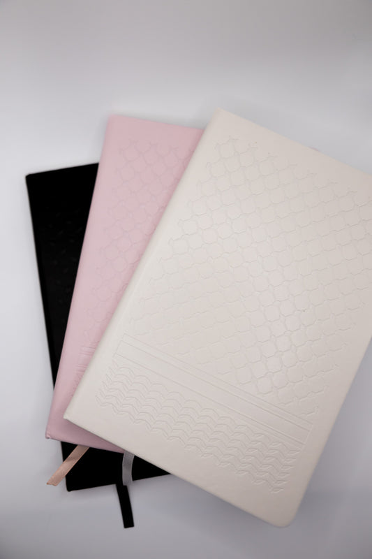 Three notebooks in black, pink, and white with a keffiyeh print embossed on the cover on a light background