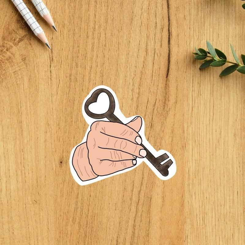 Sticker of a hand holding a key on a wooden surface with pencils and leaves.