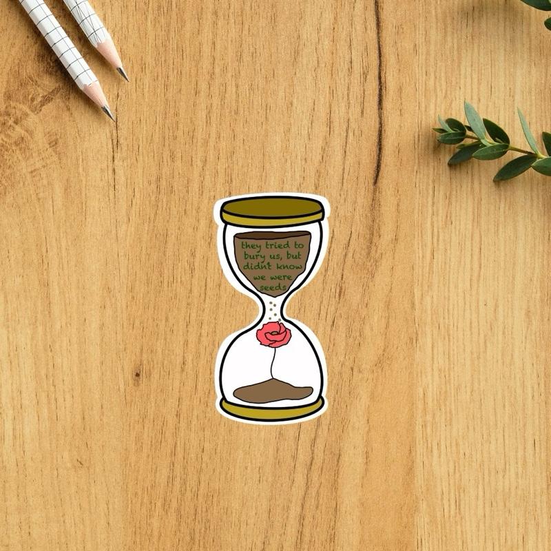 Sticker of an hourglass with a poppy flower on a wooden surface with pencils and leaves.
