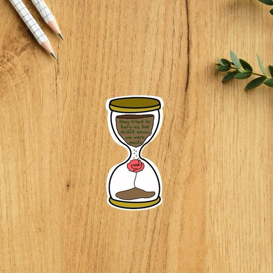 Sticker of an hourglass with a poppy flower on a wooden surface with pencils and leaves.