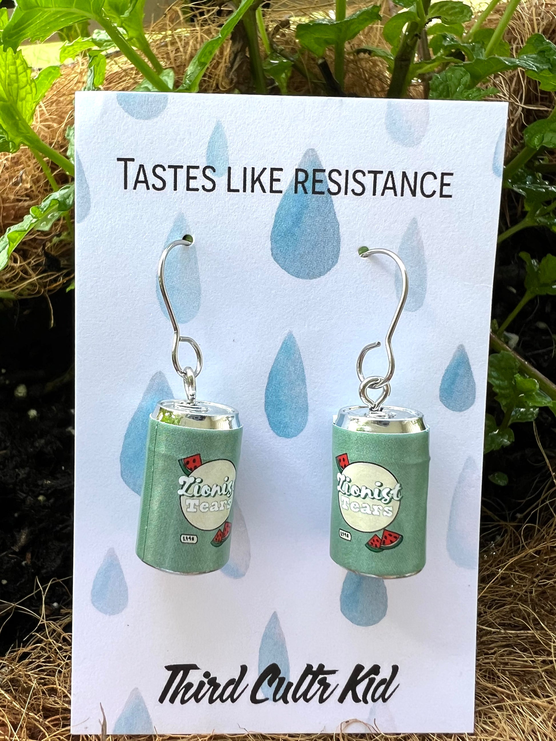 Earrings resembling a can with 'Third Cultr Kid' branding on a card with 'Tastes Like Resistance' text.