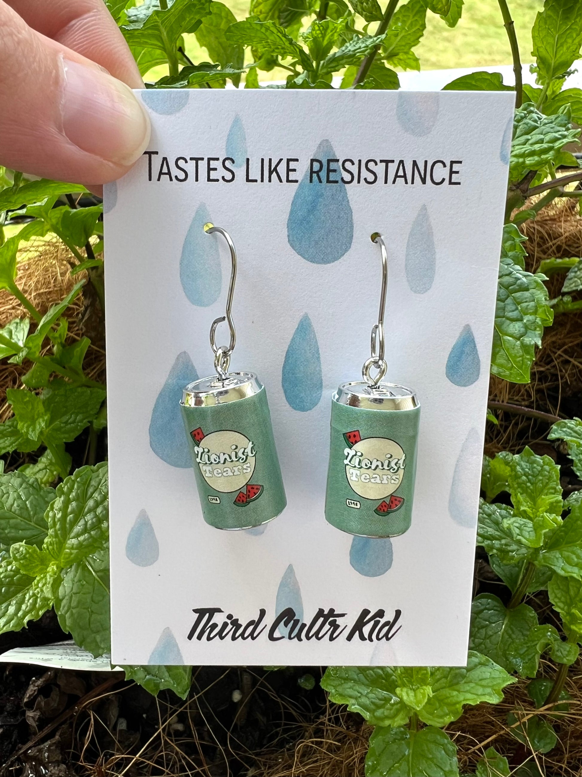 Earrings resembling green soda cans on a card with text 'Tastes Like Resistance' and 'Third Cultr Kid' against a natural background.