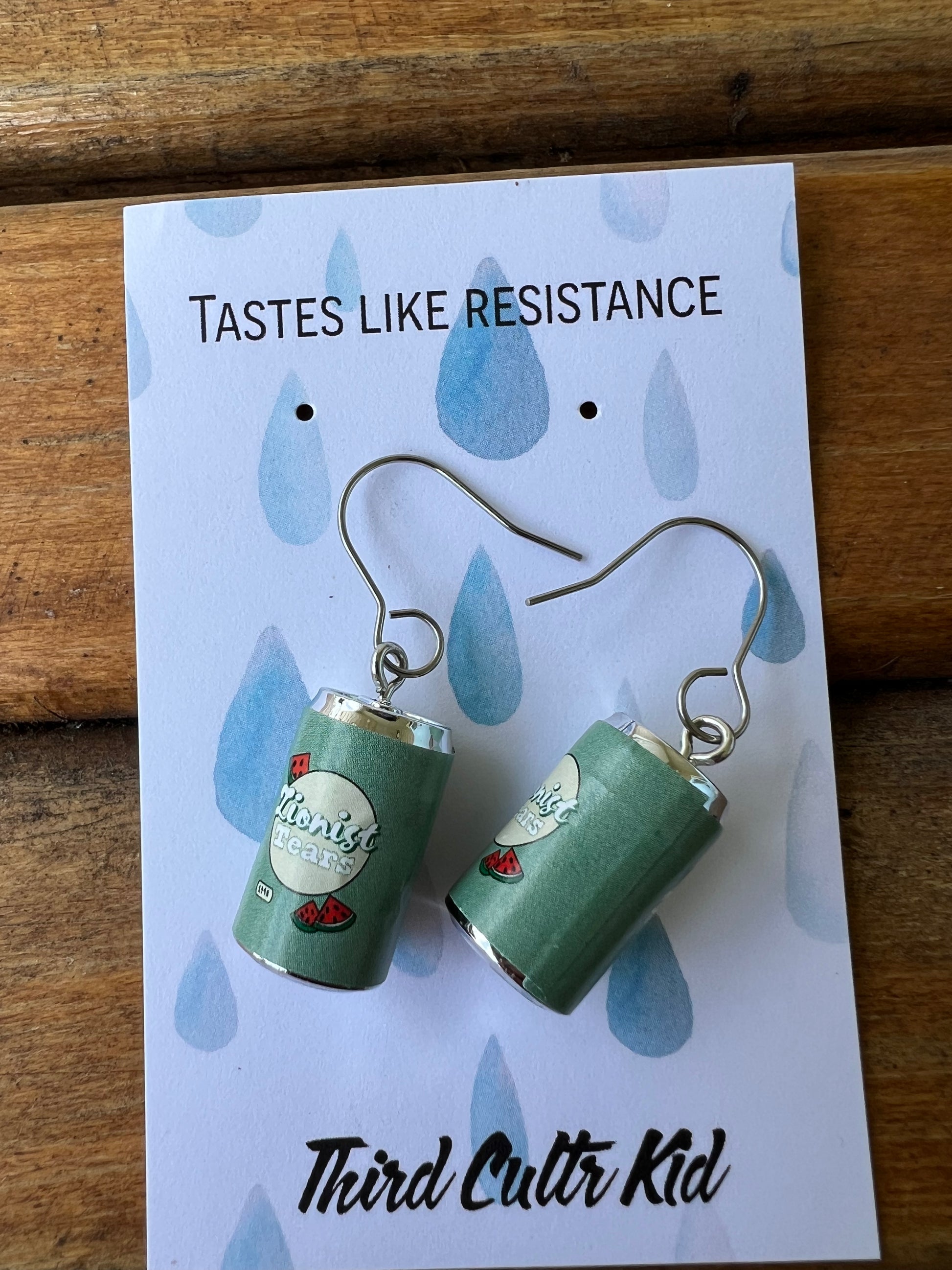 Green can-shaped earrings on a 'Tastes Like Resistance' card with 'Third Cultr Kid' branding.