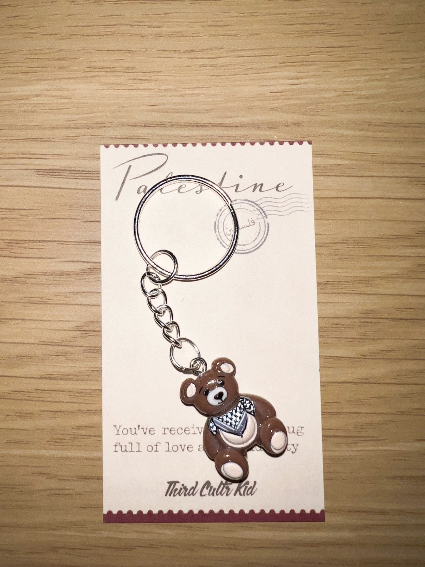 Keychain with teddy bear design on a white card with text, placed on a wooden surface.