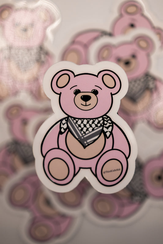 Pink teddy bear sticker with a black and white keffiyeh scarf on a blurred background of similar stickers.