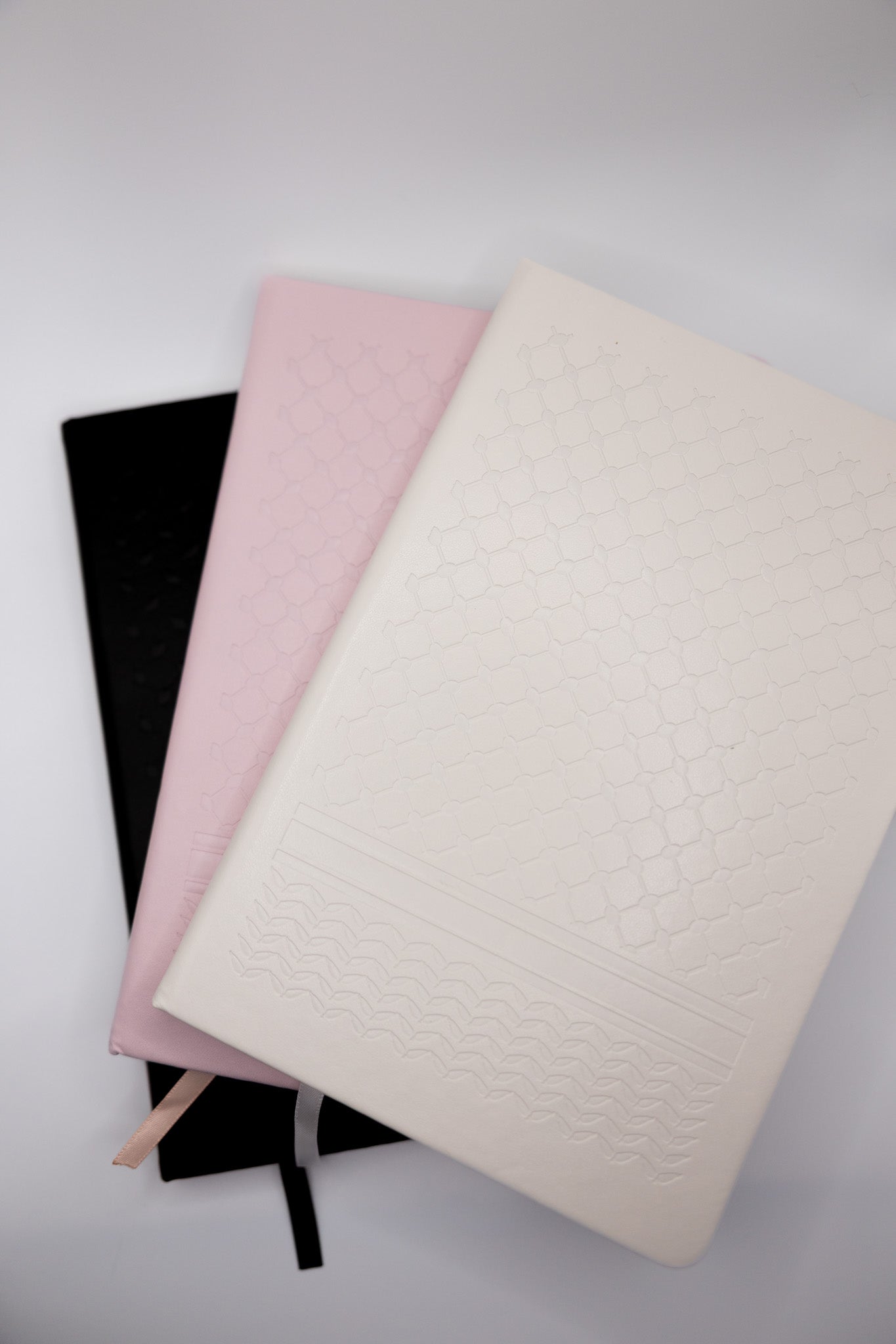 Three notebooks in black, pink, and white with a keffiyeh print embossed on the cover on a light background