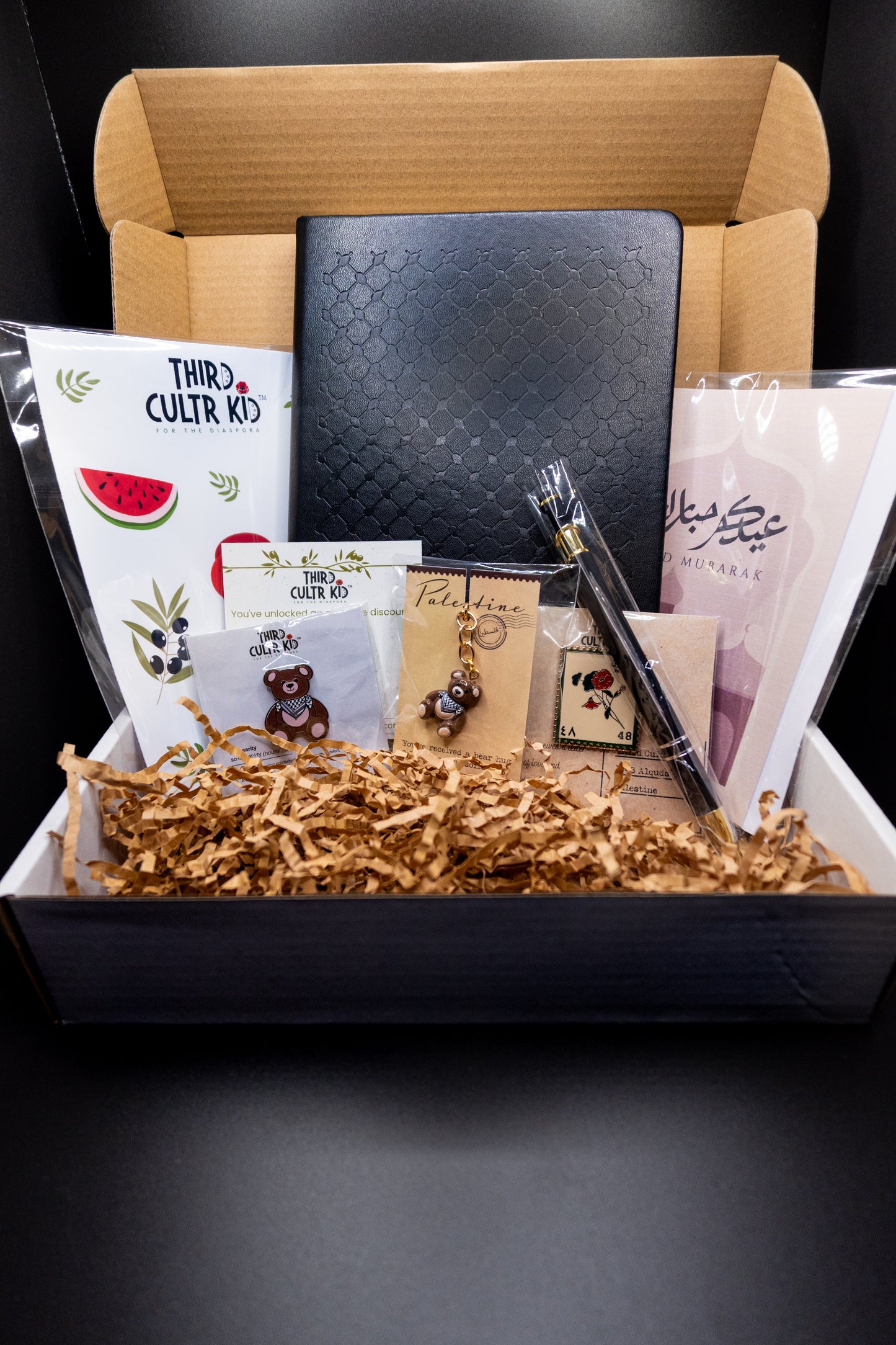 Eid and Ramadan gift box with stationery items including a keffiyeh notebook, pen, enamel pins, sticker sheet, greeting cards on a dark background