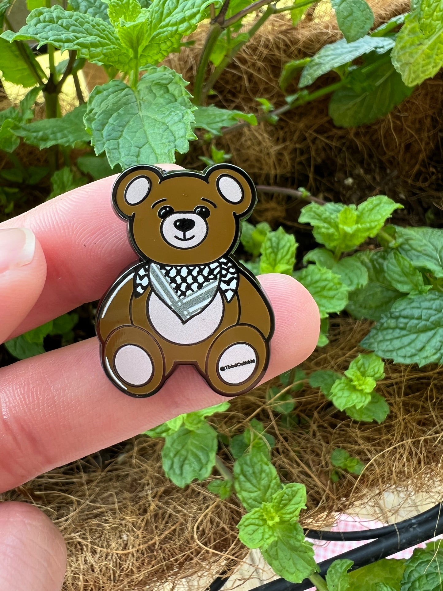 Cartoon bear pin held between fingers with green plants in the background